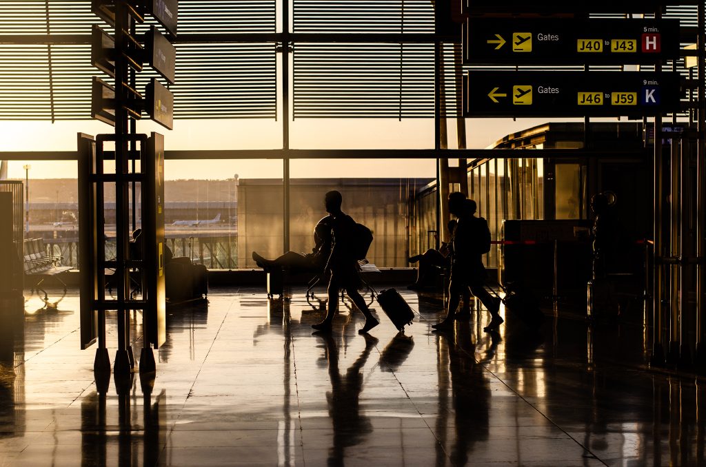 Silhouetted travelers walking near airport gates, with digital signs for J40–J59 and “5 min.” displayed.
