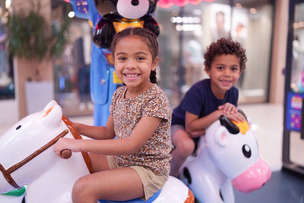 Two young children smiling while riding a colourful amusement carousel horse indoors.